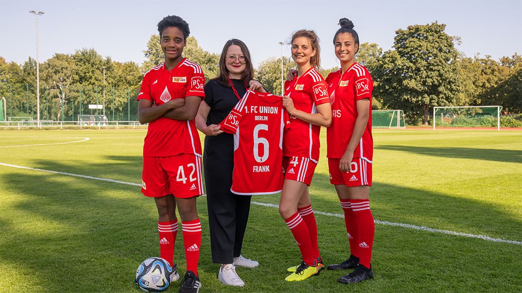 Vier Frauen in roten Fußballtrikots stehen auf einem Platz und halten ein Trikot mit der Nummer 6.
