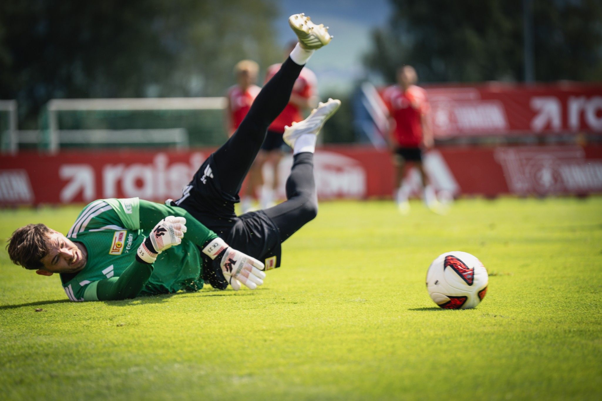 Ein Torwart in grüner Kleidung liegt auf dem Rasen, während er nach einem Ball greift. Im Hintergrund sind Spieler zu sehen.