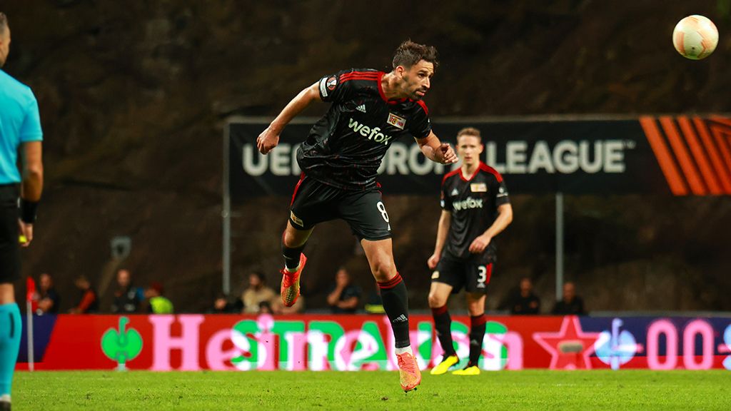 Ein Fußballspieler springt zum Kopfball, während ein Schiedsrichter und ein anderer Spieler im Hintergrund zu sehen sind.