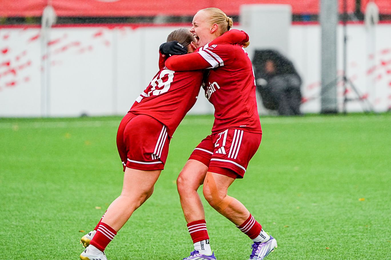 Zwei Fußballspielerinnen in roten Trikots umarmen sich freudig auf einem Trainingsplatz.
