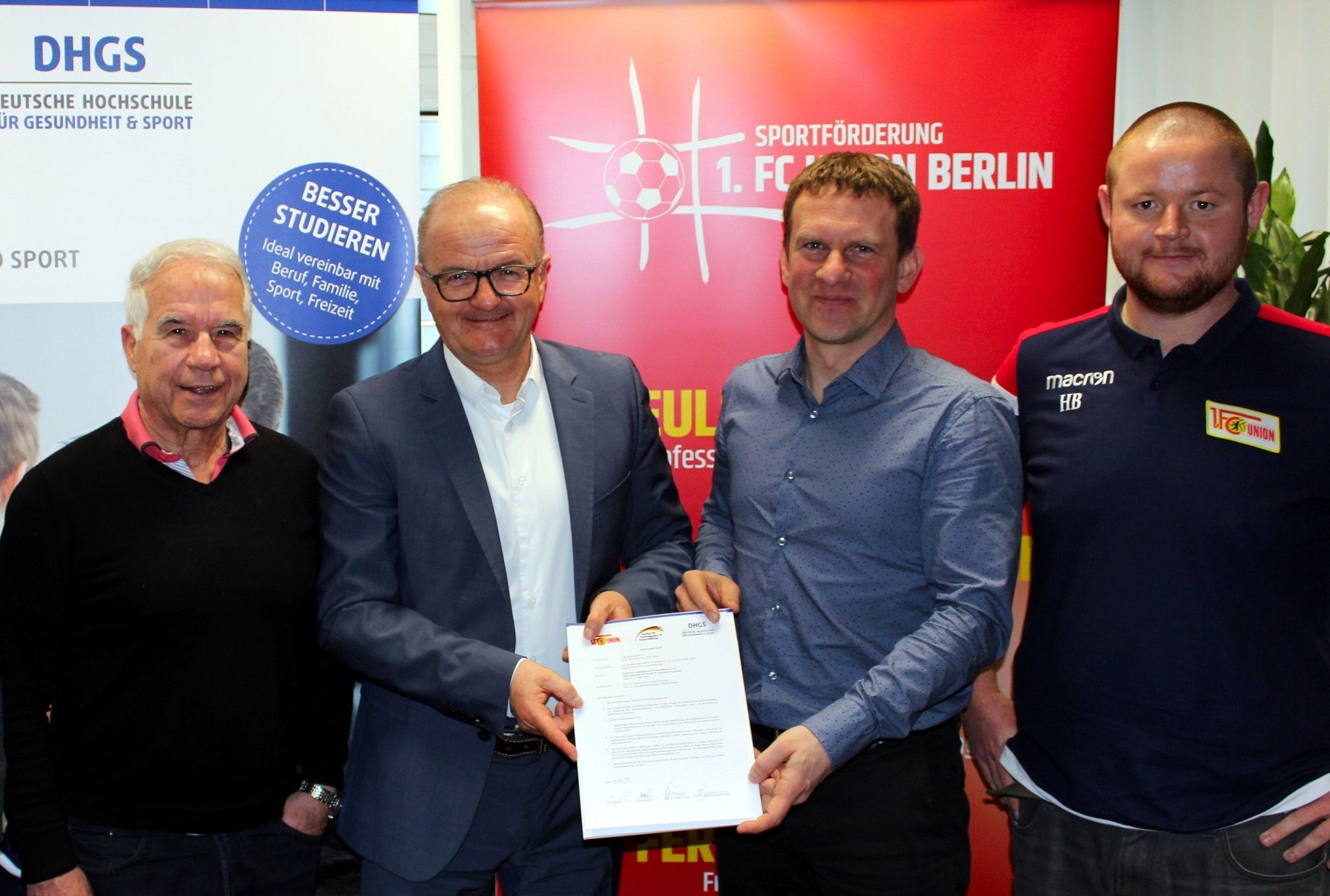 Vier Männer stehen zusammen und halten ein Dokument, mit Logos im Hintergrund, das eine Zusammenarbeit symbolisiert.