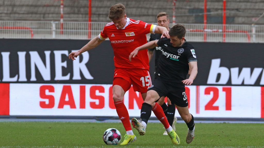 A soccer player in a red jersey dribbles the ball while a player in a dark jersey tries to stop him.