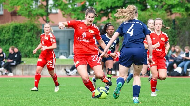 Zwei Frauenfußballmannschaften spielen auf einem Rasenfeld; eine Spielerin in Rot dribbelt den Ball, während eine Gegnerin sie verteidigt.