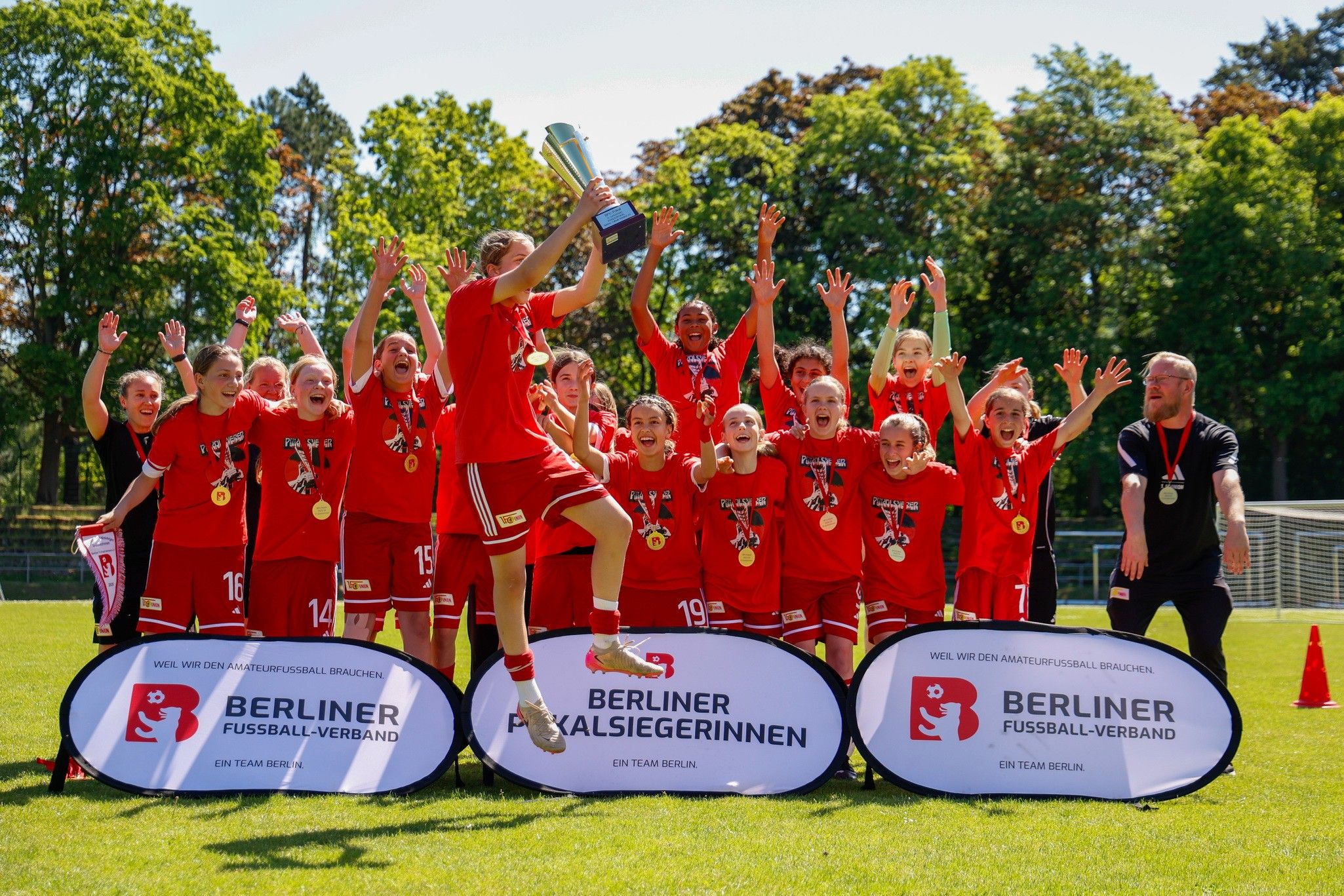 Ein jubelndes Fußballteam in roten Trikots feiert den Sieg mit Pokal und Medaillen auf einem Sportplatz.