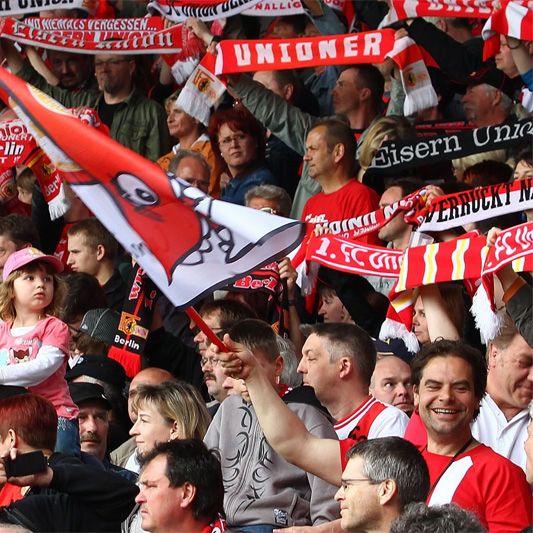 Eine Menschenmenge in einem Stadion, die enthusiastisch Teamfahnen und Schals hält und Freude zeigt.