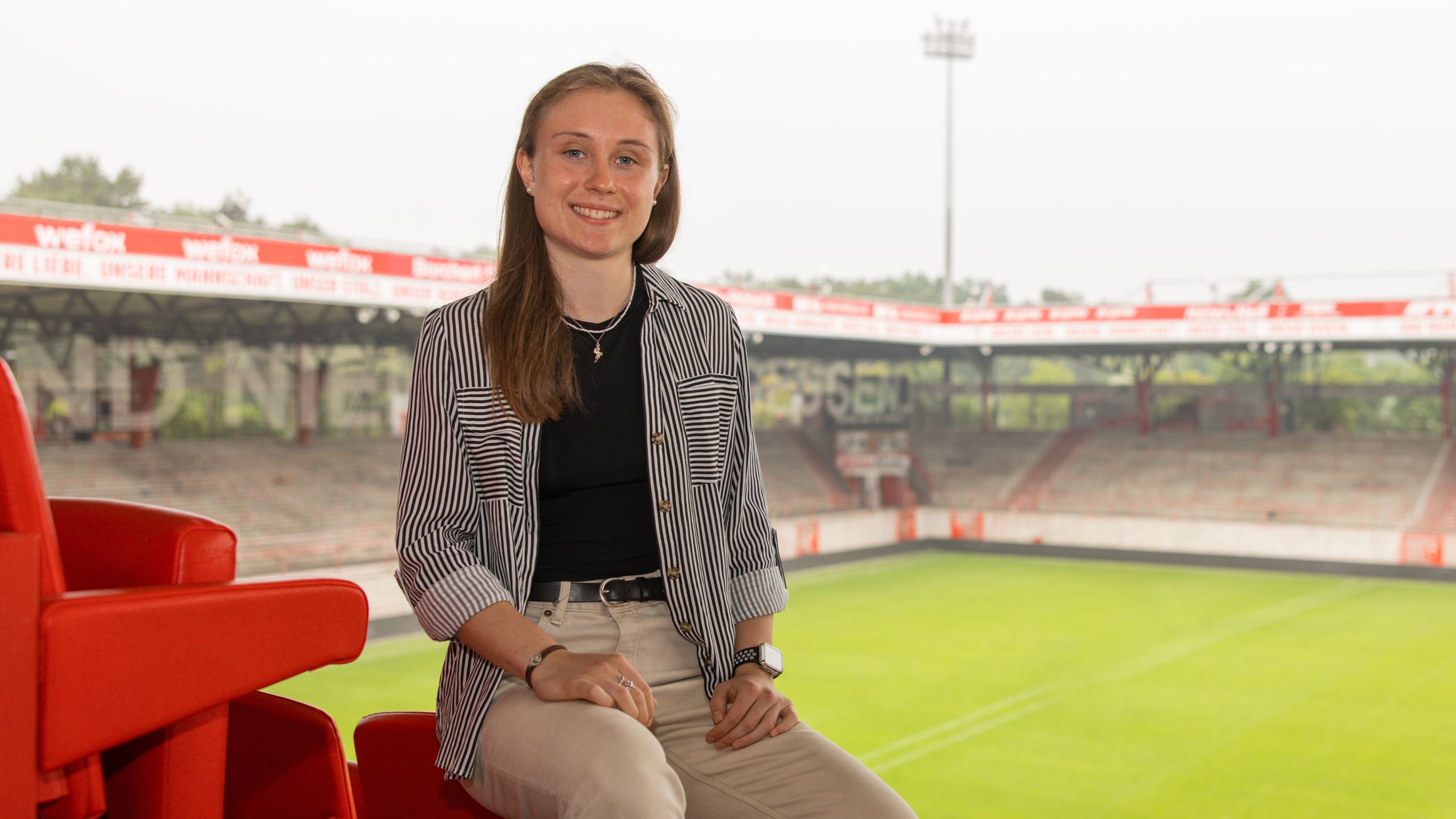 A young woman sits on a red seating area in the stadium, with a green playing field in the background.