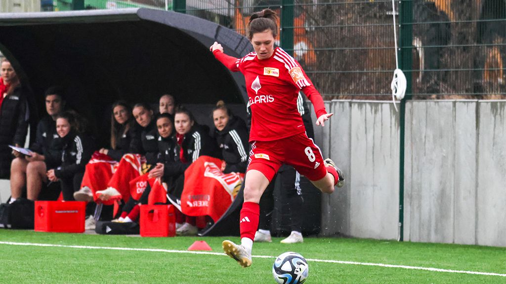 Eine Sportlerin in rotem Trikot schießt einen Fußball auf einem Trainingsplatz, umgeben von anderen Spielerinnen und Trainerinnen.