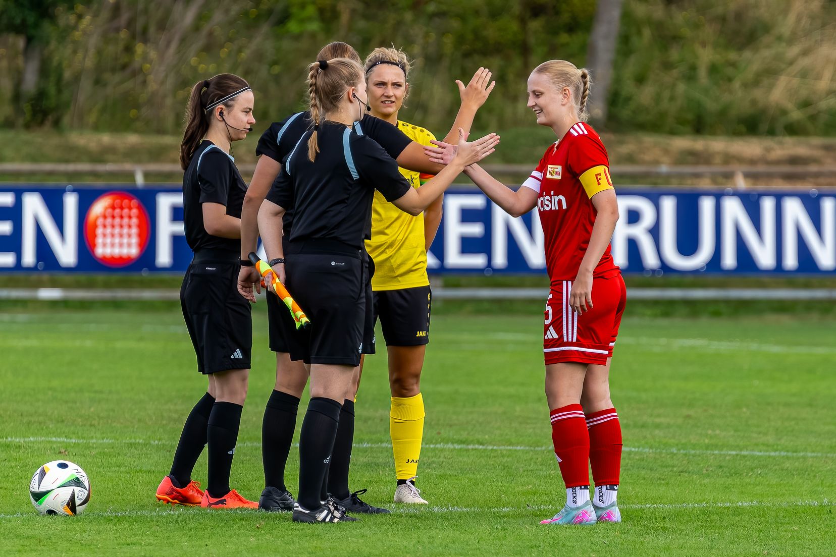 Vier Fußballspielerinnen und eine Schiedsrichterin stehen auf dem Spielfeld, schütteln sich die Hände und lächeln.