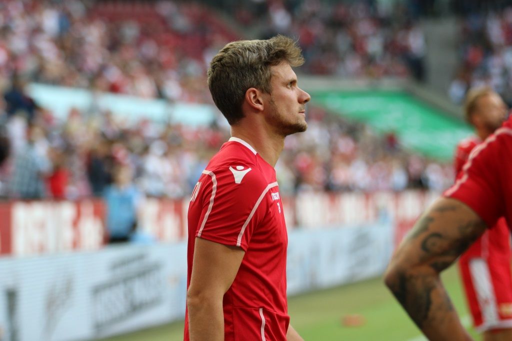 Ein Fußballspieler in einem roten Trikot steht an der Seitenlinie und schaut nachdenklich in die Ferne.