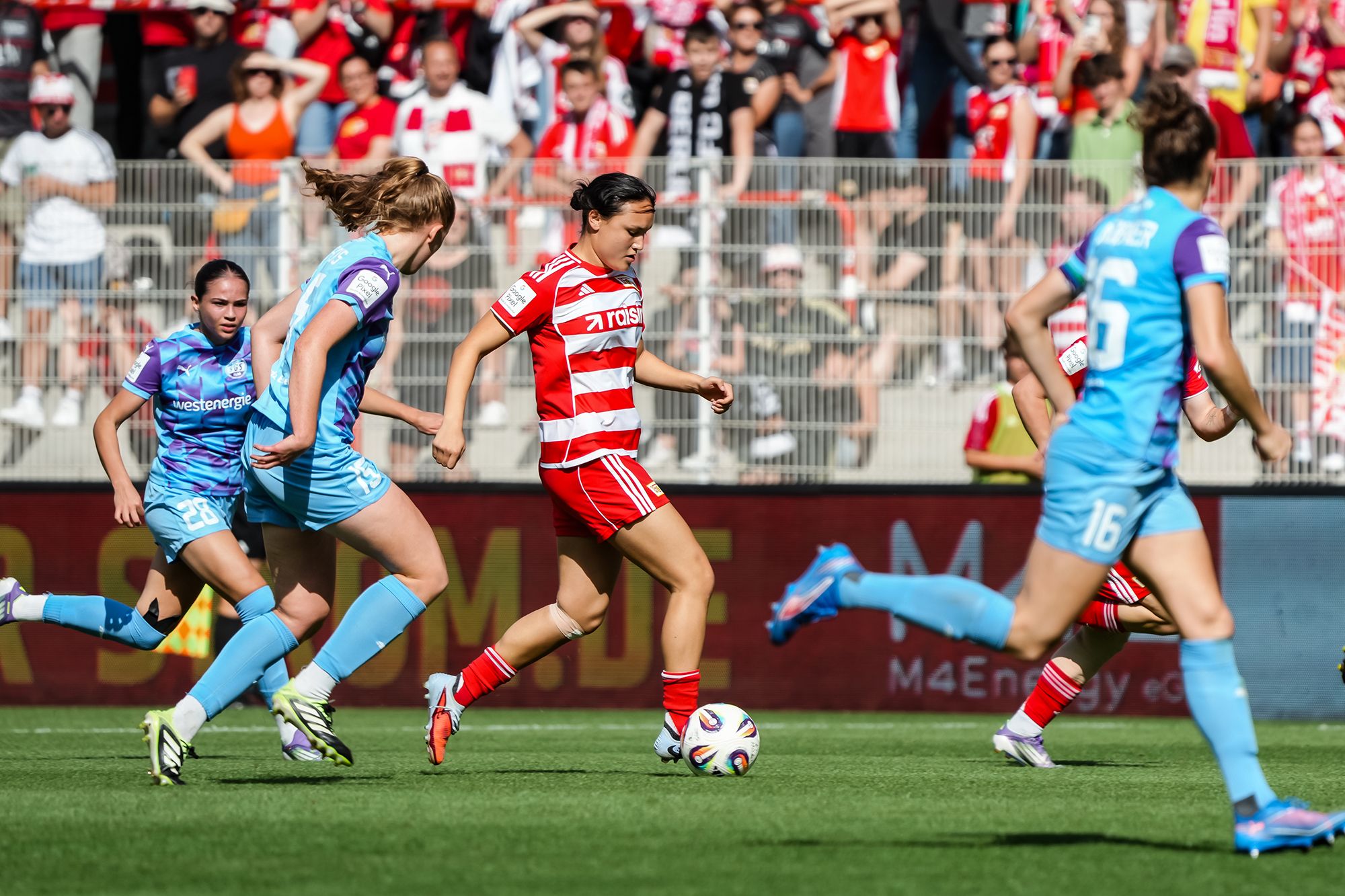 Eine Fußballspielerin in rot-weißem Trikot dribbelt den Ball, während mehrere Gegnerinnen in blauen Trikots angreifen.
