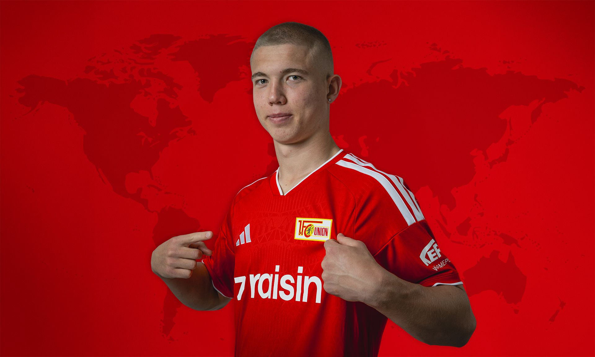 Ein sportlicher junger Mann in einem roten Fußballtrikot steht vor einer roten Weltkarte und zeigt mit den Daumen auf sich selbst.