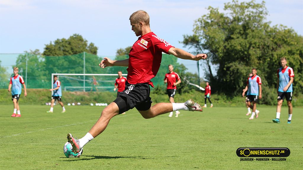 Fussballspieler in rotem Trikot schießt während eines Trainings auf einem grünen Platz, im Hintergrund weitere Spieler.