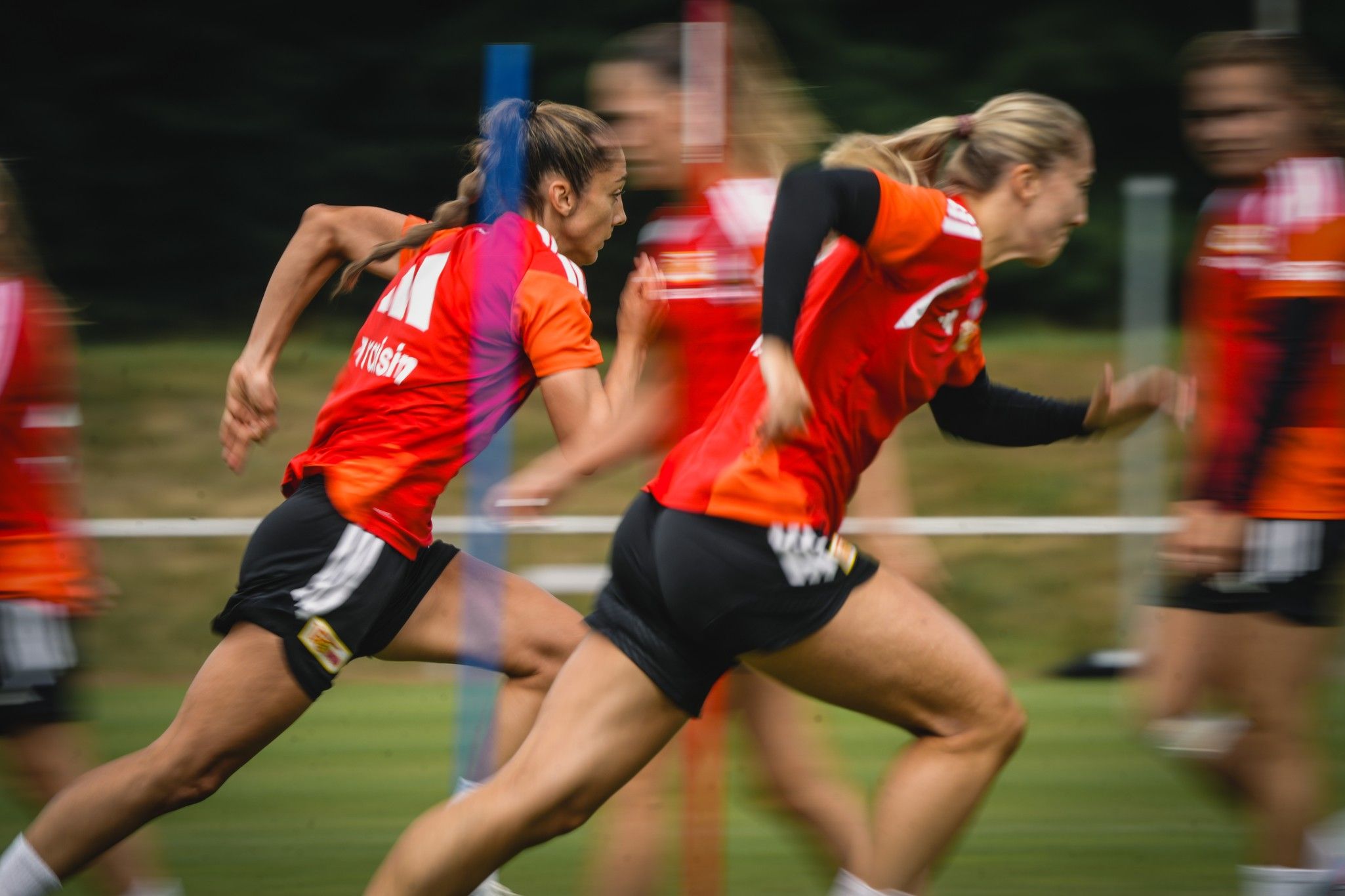 Zwei Frauen in roten Trikots sprinten schnell auf einem Trainingsfeld, während andere Spieler im Hintergrund unscharf sind.