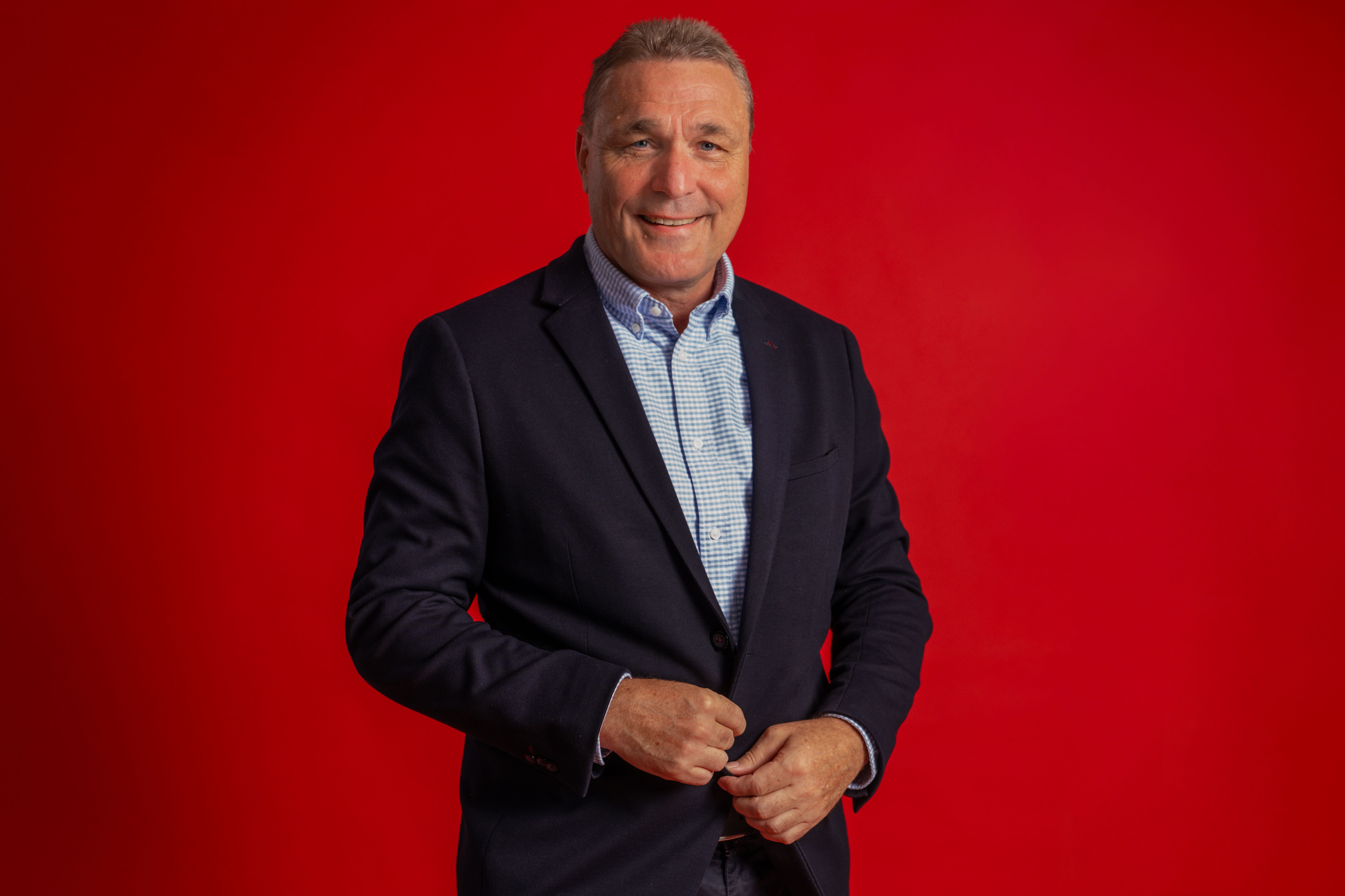 Mann in dunkelblazer und kariertem Hemd, lächelt vor rotem Hintergrund.