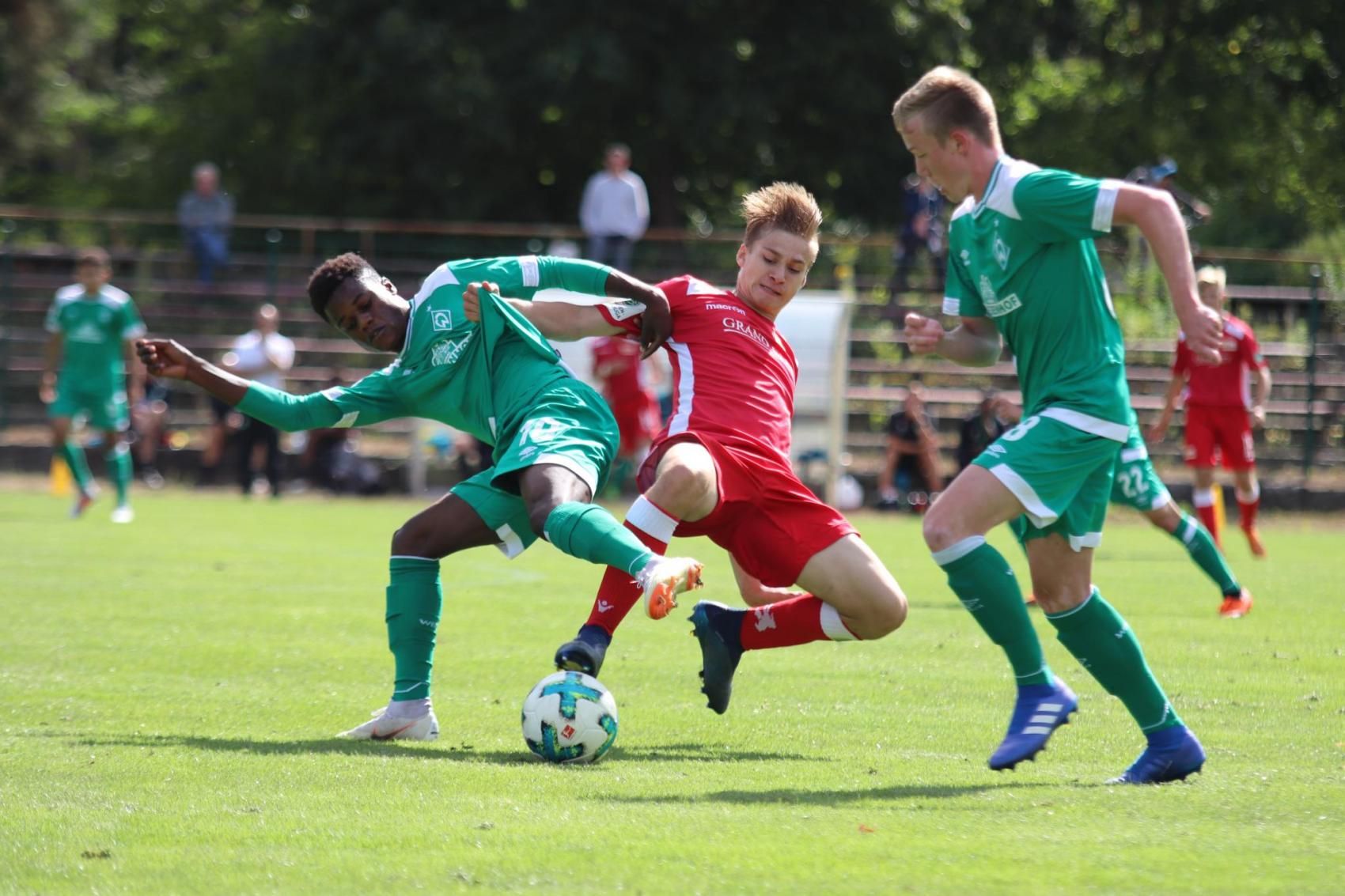 Zwei Fußballspieler in grünen Trikots im Zweikampf mit einem Spieler in einem roten Trikot auf einem Rasenfeld.