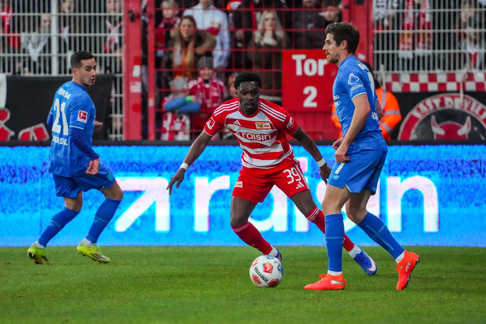 Ein Fußballspieler in roter und weißer Uniform dribbelt den Ball, während zwei gegnerische Spieler in blauen Trikots ihn umgeben.