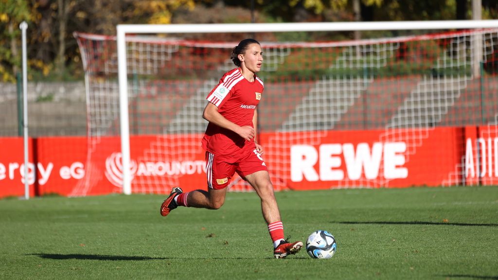 Ein Fußballspieler in roter Ausrüstung läuft mit dem Ball auf einem Trainingsplatz. Im Hintergrund sind Tore und Werbebanner sichtbar.