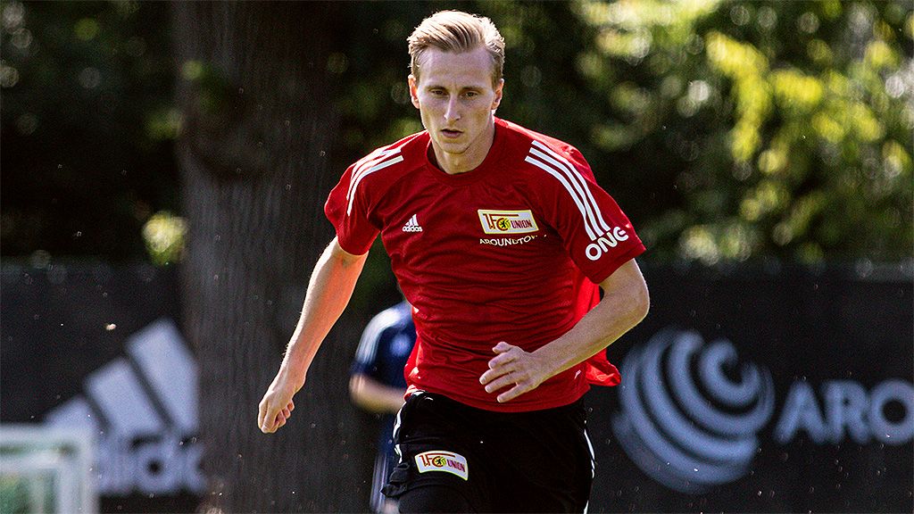 Ein Fußballspieler in einem roten Trikot sprintet auf einem Trainingsfeld unter Bäumen.