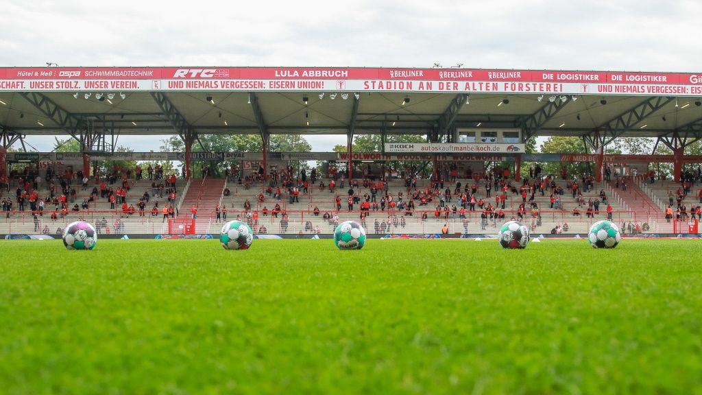 Fünf Fußballbälle auf grünem Rasen vor einer Zuschauertribüne in einem Stadion, das teilweise leer ist.