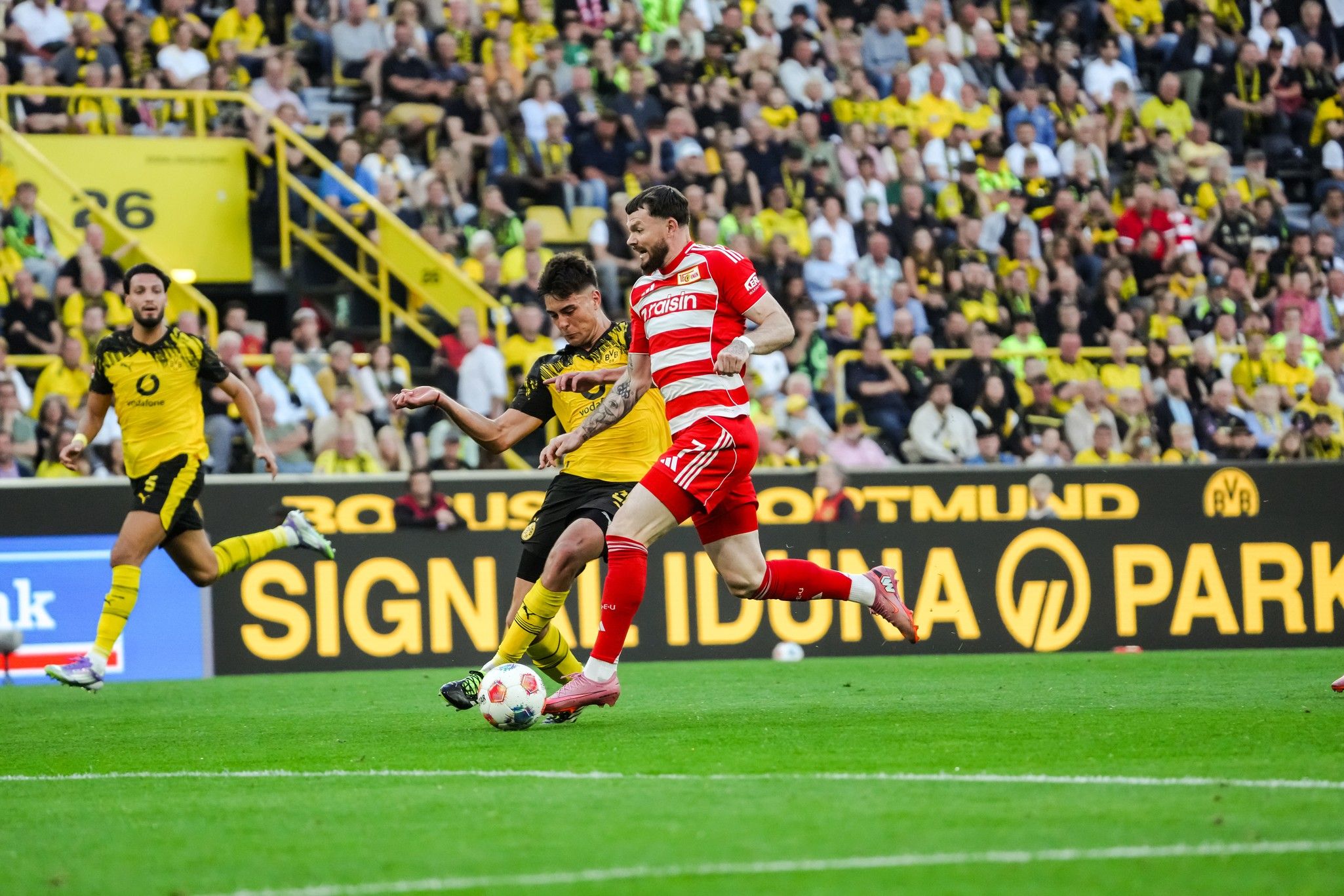 Ein Fußballspieler in gelber Ausrüstung wird von einem Spieler in roter Ausrüstung im Spiel am Ball angegriffen. Zuschauer im Hintergrund.