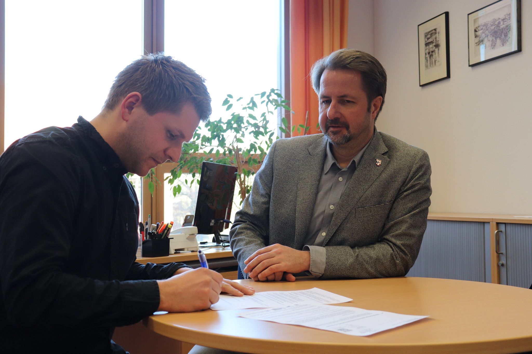 Ein Mann sitzt an einem Tisch und unterschreibt Dokumente, während ein weiterer Mann daneben aufmerksam zuschaut.