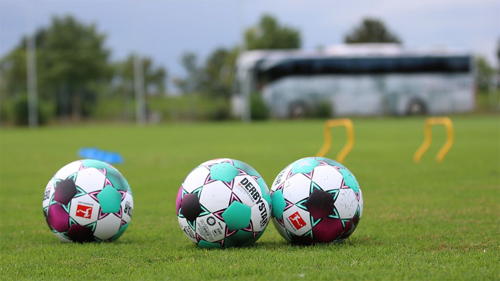 Drei Fußball mit bunten Mustern liegen auf einem grünen Platz, im Hintergrund ist ein Bus sichtbar.