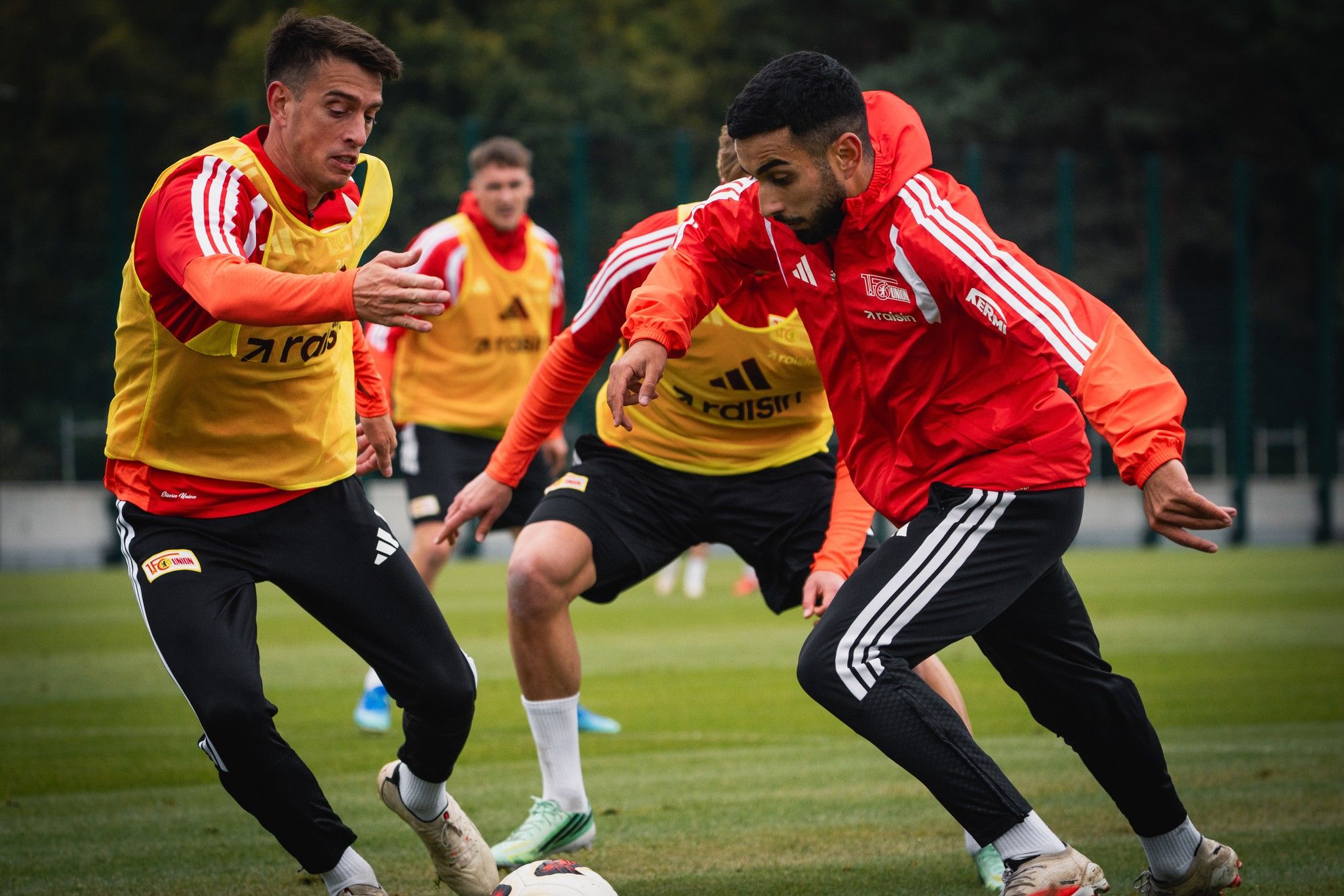 Zwei Fußballspieler im Training dribbeln den Ball, während andere im Hintergrund Übungen machen. Alle tragen Trainingskleidung.