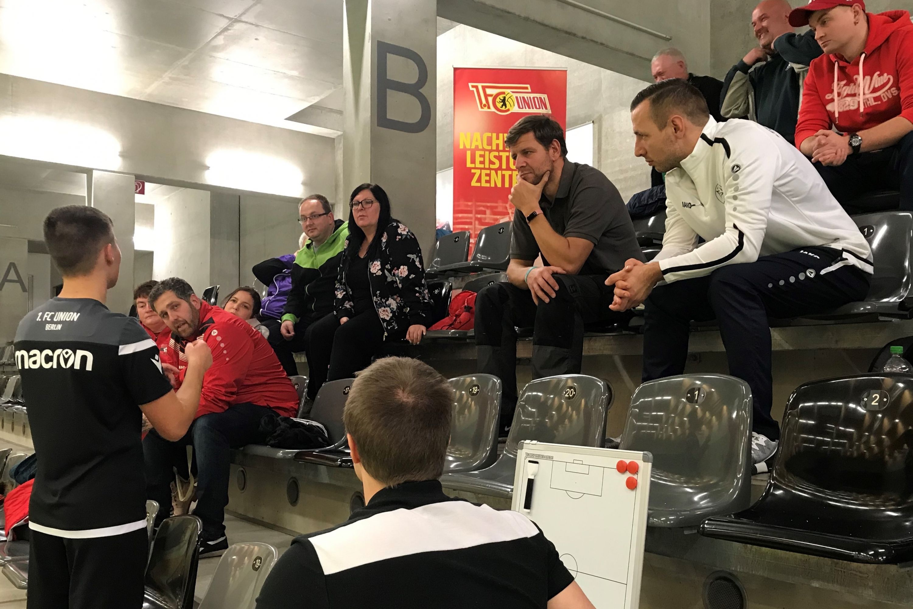 Eine Gruppe von Menschen sitzt auf Tribünen und beobachtet eine Besprechung. Im Vordergrund steht ein Trainer mit einem Taktikboard.