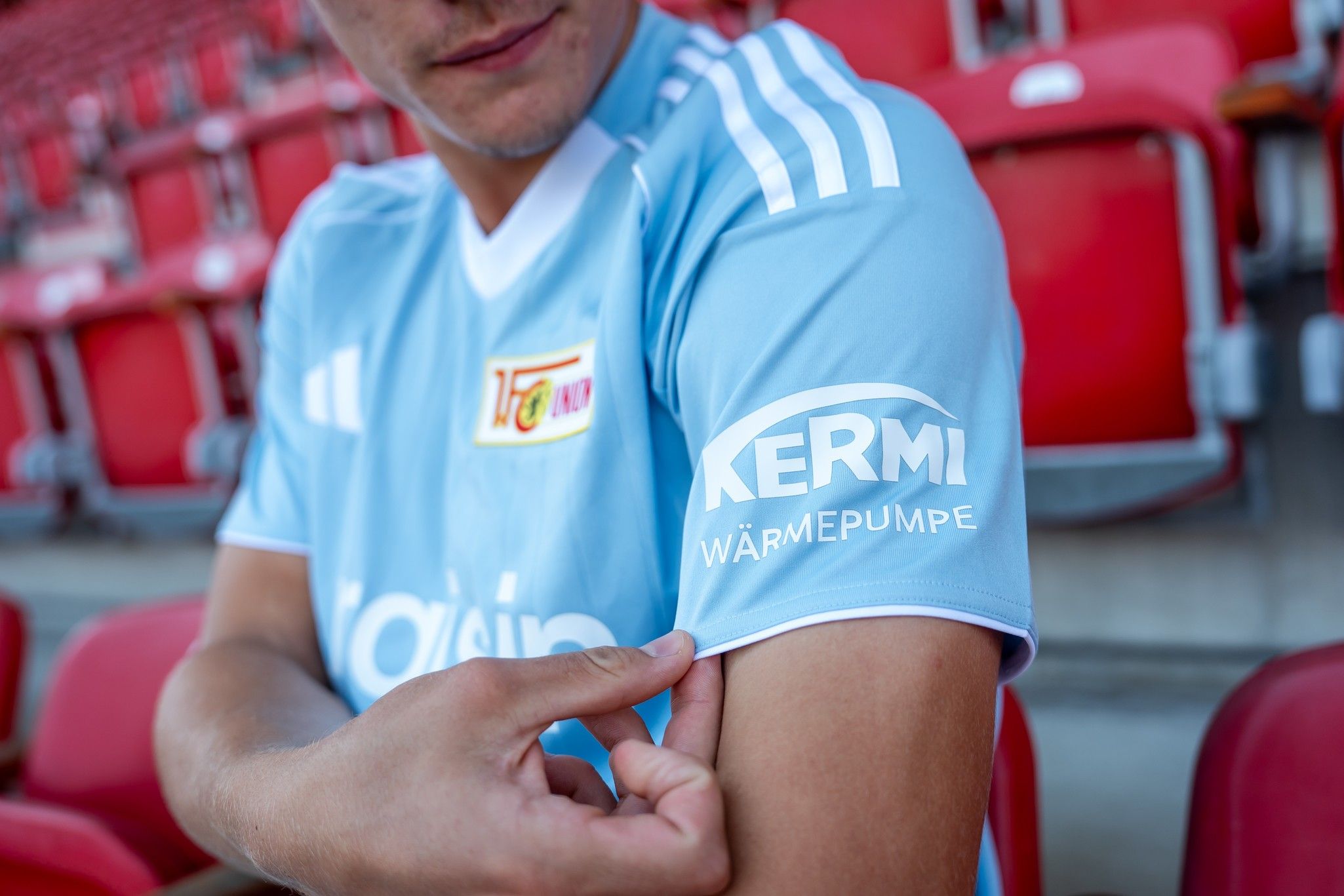 Ein sportlich gekleideter Mann zeigt seinen Arm in einem hellblauen Trikot mit dem Logo "Kermi Wärmepumpe" auf dem Ärmel.