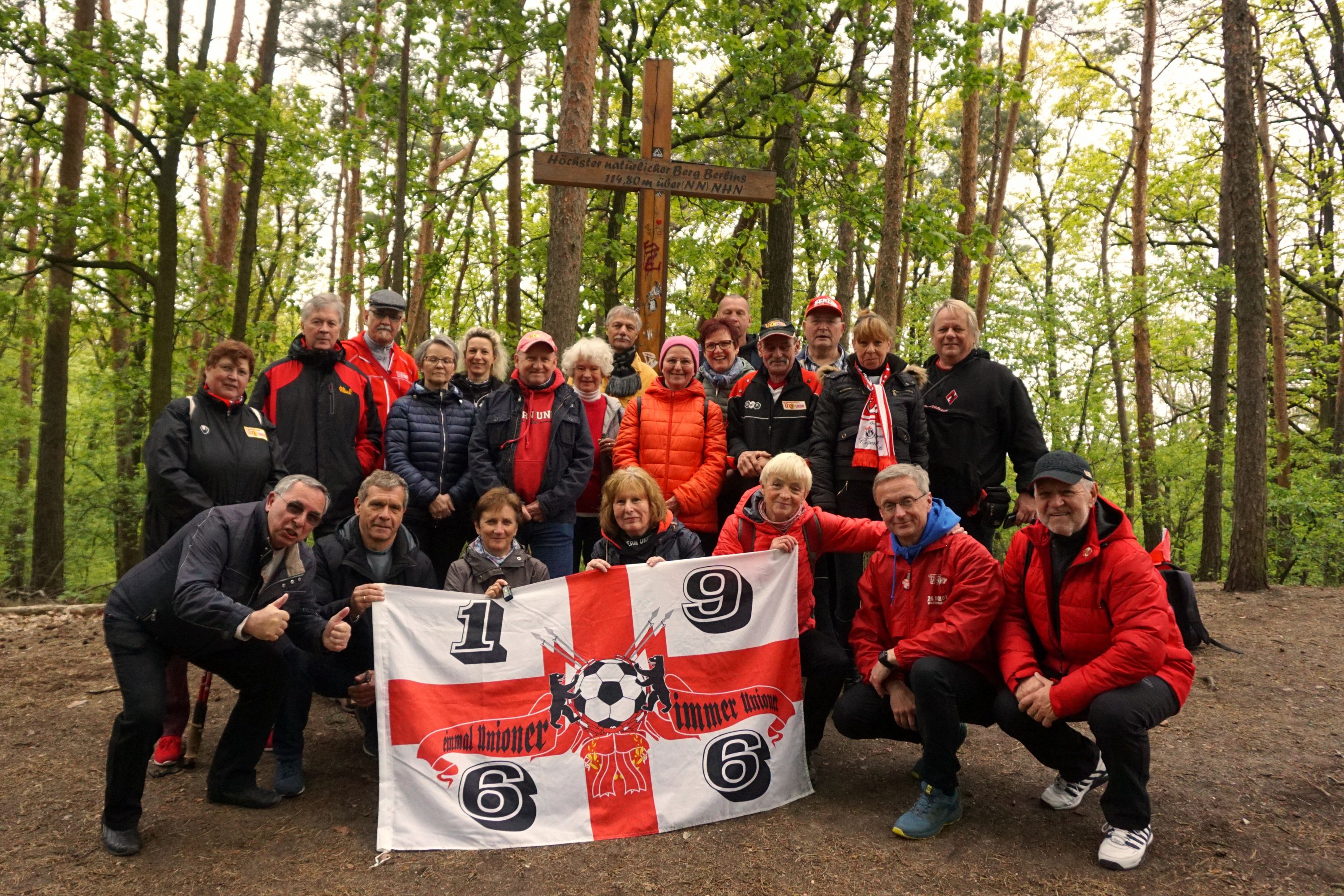 Gruppe von Menschen in roten Jacken, die ein Fußballbanner halten, umgeben von Bäumen in einem Waldgebiet.