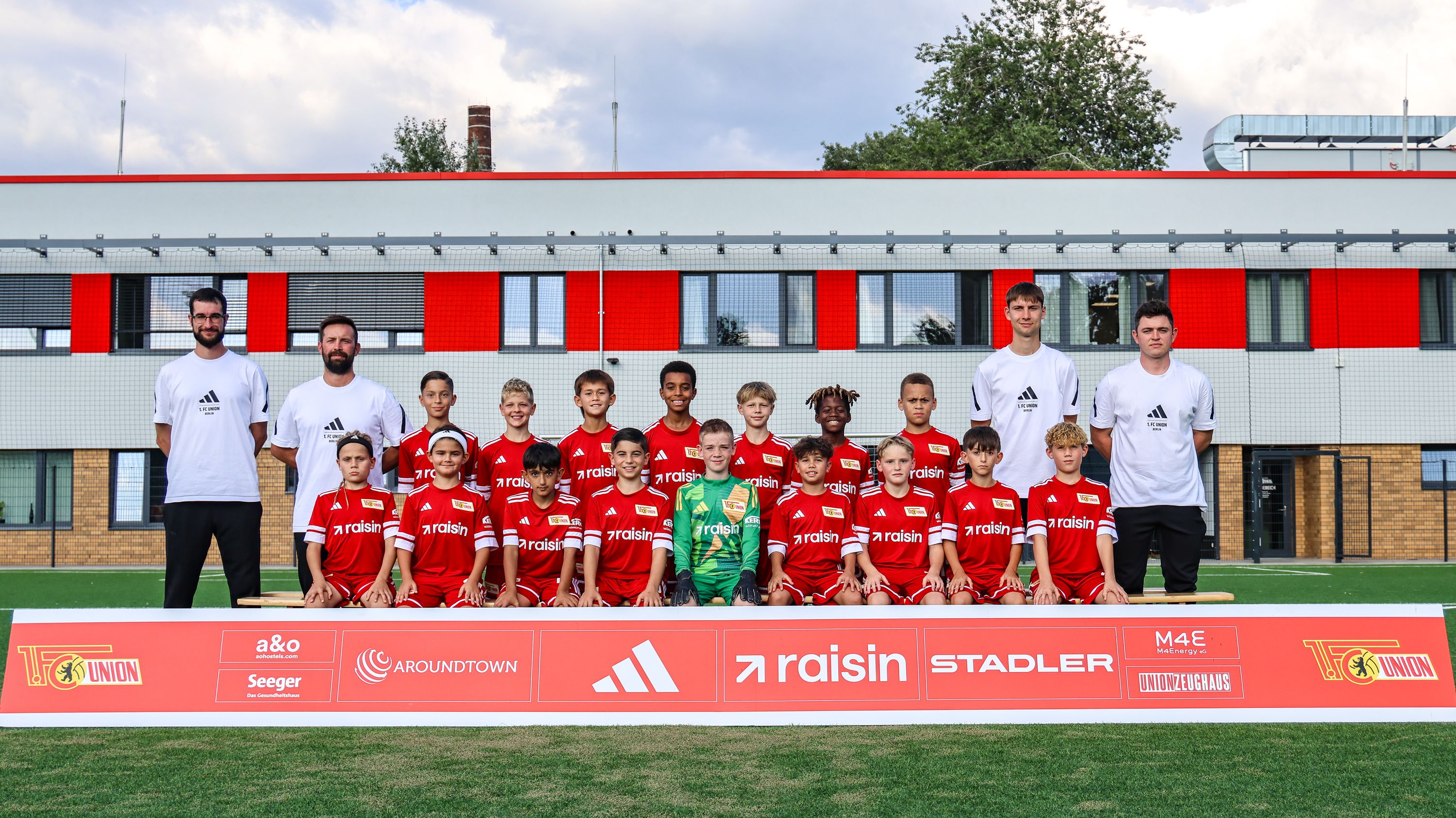 Mannschaftsfoto einer Fußballjugendmannschaft in roten Trikots auf einem Sportplatz, umgeben von Trainern.