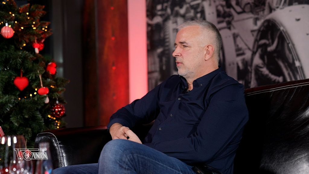 Mann mit grauen Haaren sitzt auf einem Sofa, im Hintergrund ein Weihnachtsbaum mit roten Dekorationen.