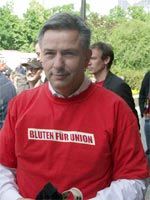 Mann in rotem T-Shirt mit der Aufschrift "BLUTEN FÜR UNION" steht vor einer Gruppe von Menschen im Freien.
