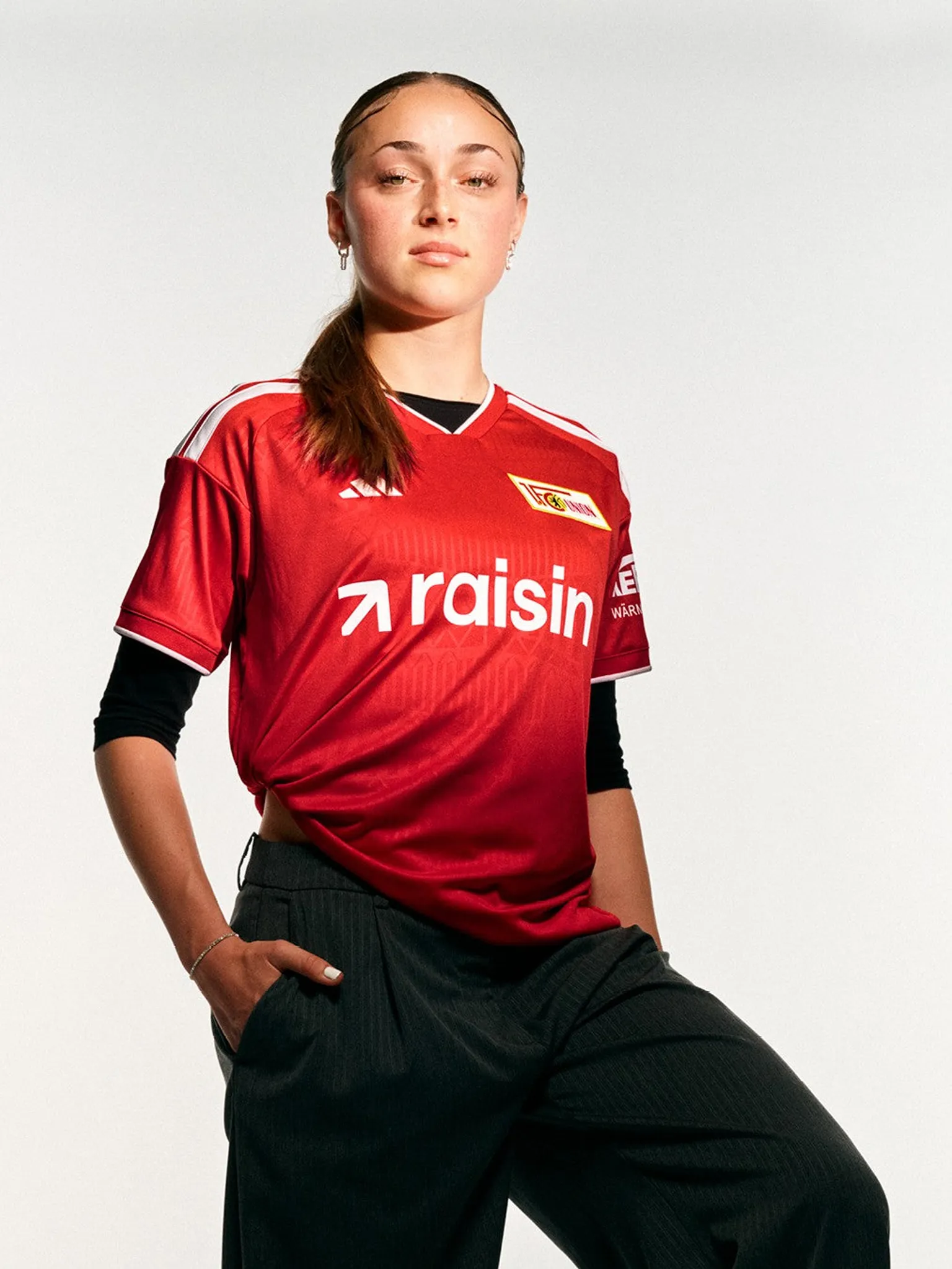 Frau in rotem Fußballtrikot mit dem Schriftzug „raisin“ und schwarzer Hose, steht selbstbewusst vor neutralem Hintergrund.