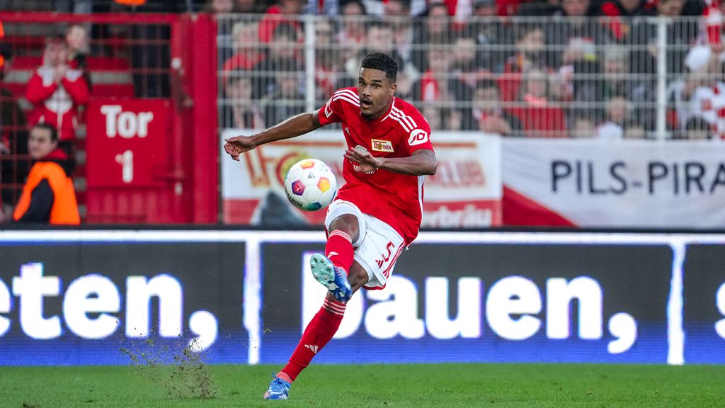 Ein Fußballspieler in roter Uniform schießt den Ball während eines Spiels, im Hintergrund jubelnde Fans.