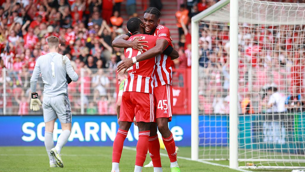 Two soccer players in red and white jerseys embrace on the field, while the goalkeeper stands in the background.