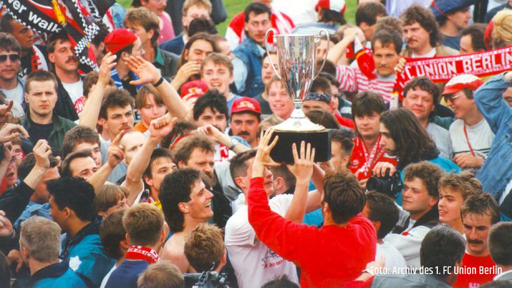 Menschenmenge feiert um einen großen Pokal, jubelnde Gesichter und Vereinsfarben sichtbar.