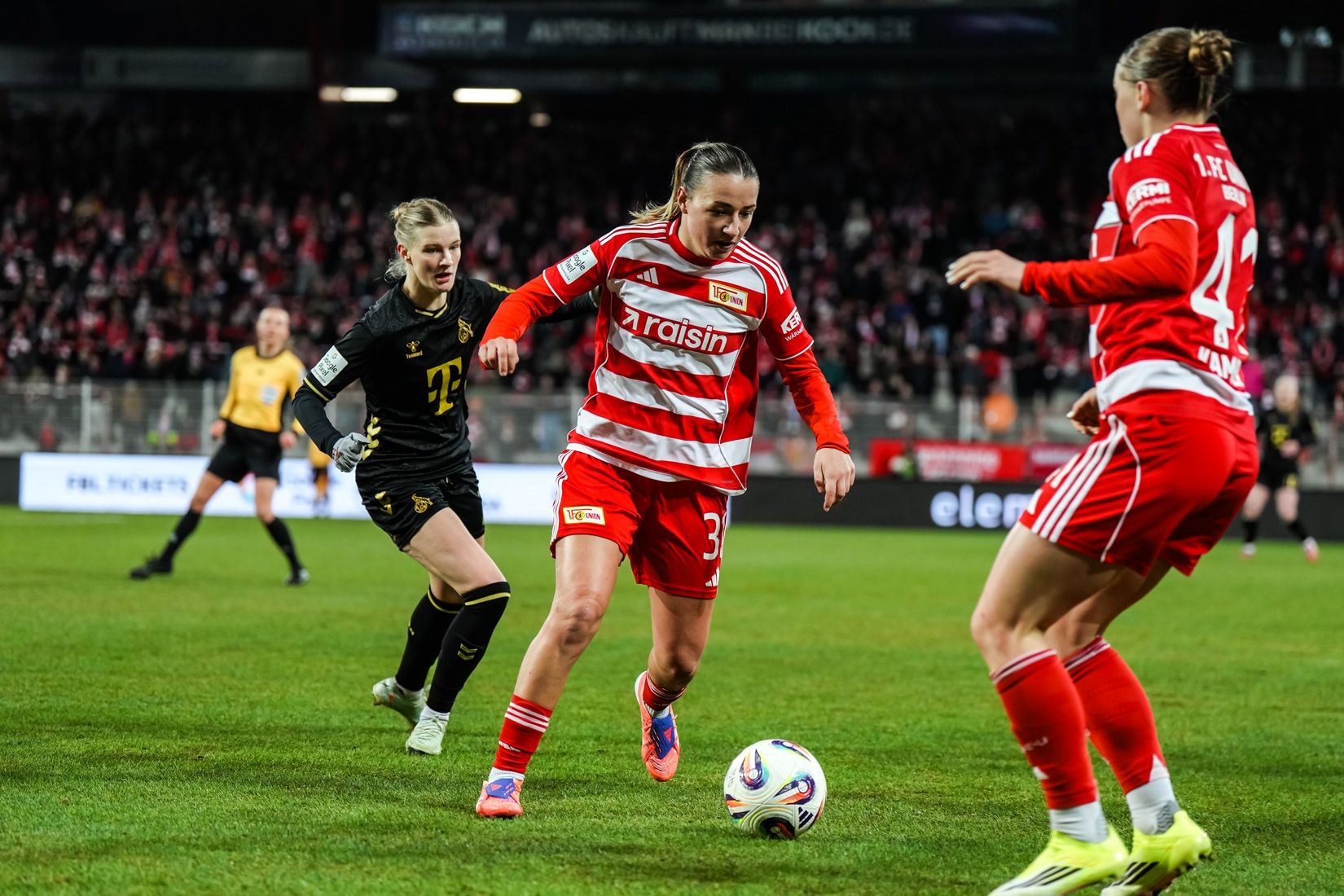 Eine Fußballspielerin in rot-weißer Uniform dribbelt den Ball, während zwei Gegnerinnen in schwarzer und gelber Kleidung sie verfolgen.