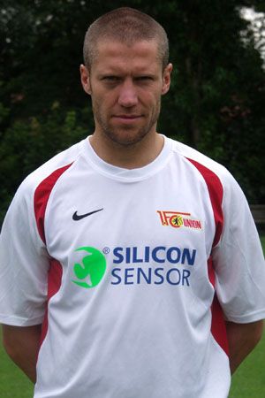 Ein Fußballspieler steht in einem weißen Team-Trikot mit roten Details und einem Sponsorenlogo auf der Vorderseite.