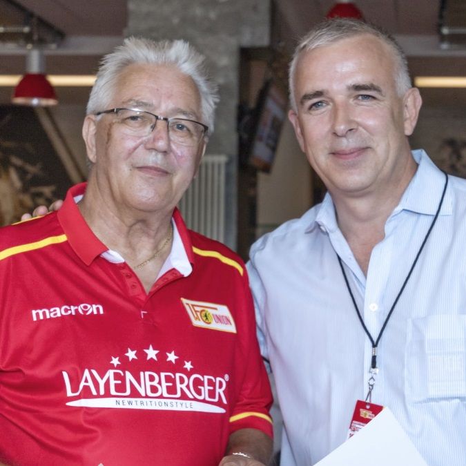 Zwei Männer lächeln in einem Restaurant, einer trägt ein rotes Fußballtrikot, der andere ein hellblaues Hemd.