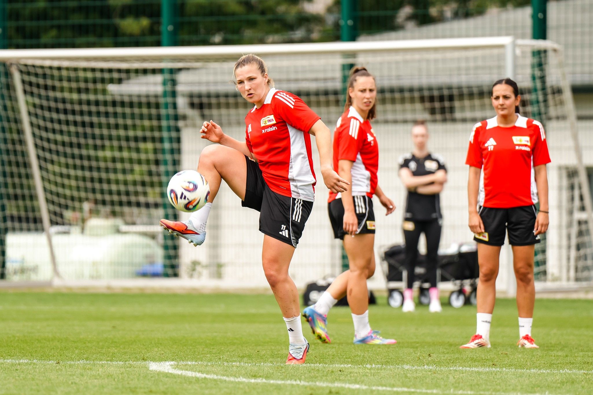 Eine Gruppe weiblicher Fußballspielerinnen trainiert auf einem Rasenplatz, während eine Spielerin den Ball mit dem Fuß kontrolliert.