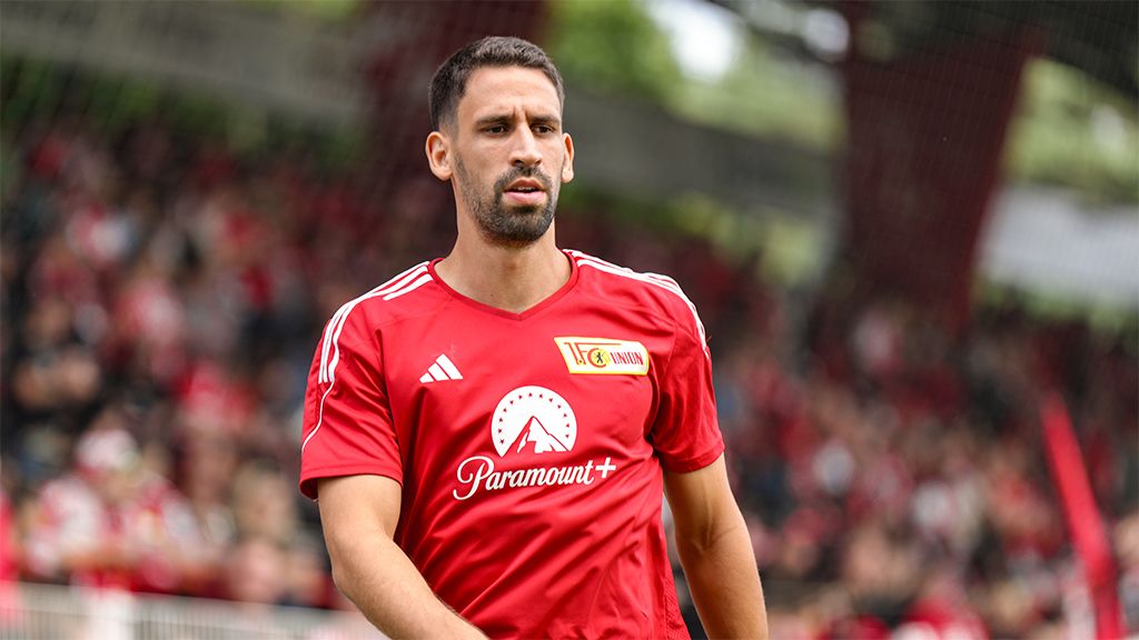 Ein Fußballspieler in rotem Trikot mit einem Vereinslogo steht auf dem Spielfeld, im Hintergrund unscharfe Zuschauer.