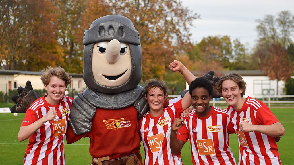 Vierte Fußballmannschaft posiert mit einem Maskottchen in Ritterkostüm auf einem Sportplatz. Alle lächeln und zeigen ihre Fäuste.