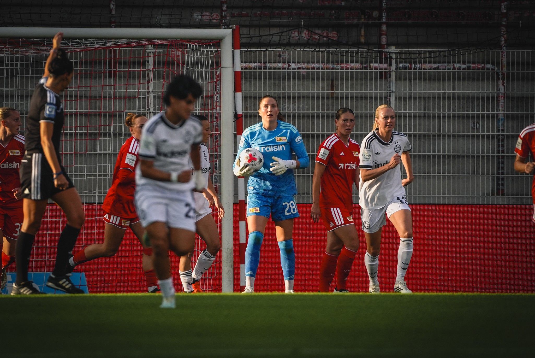 Torhüterin in blauer Kleidung steht hinter Abwehrspielerinnen, während Spielerinnen in roten Trikots sich auf einem Fußballfeld positionieren.