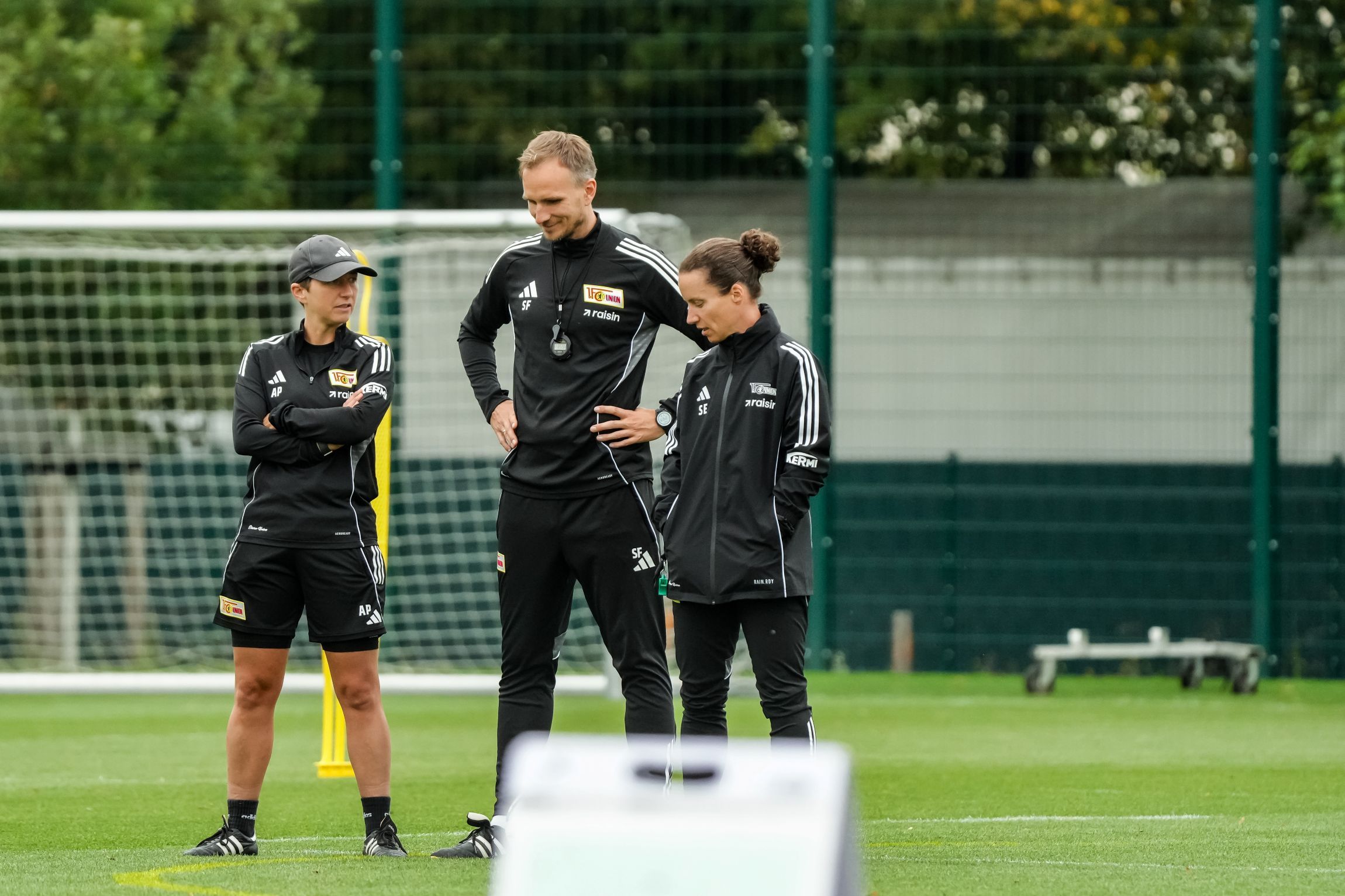 Drei Trainer auf einem Fußballplatz, einer steht in der Mitte, während die anderen beiden gebannt auf den Boden schauen.