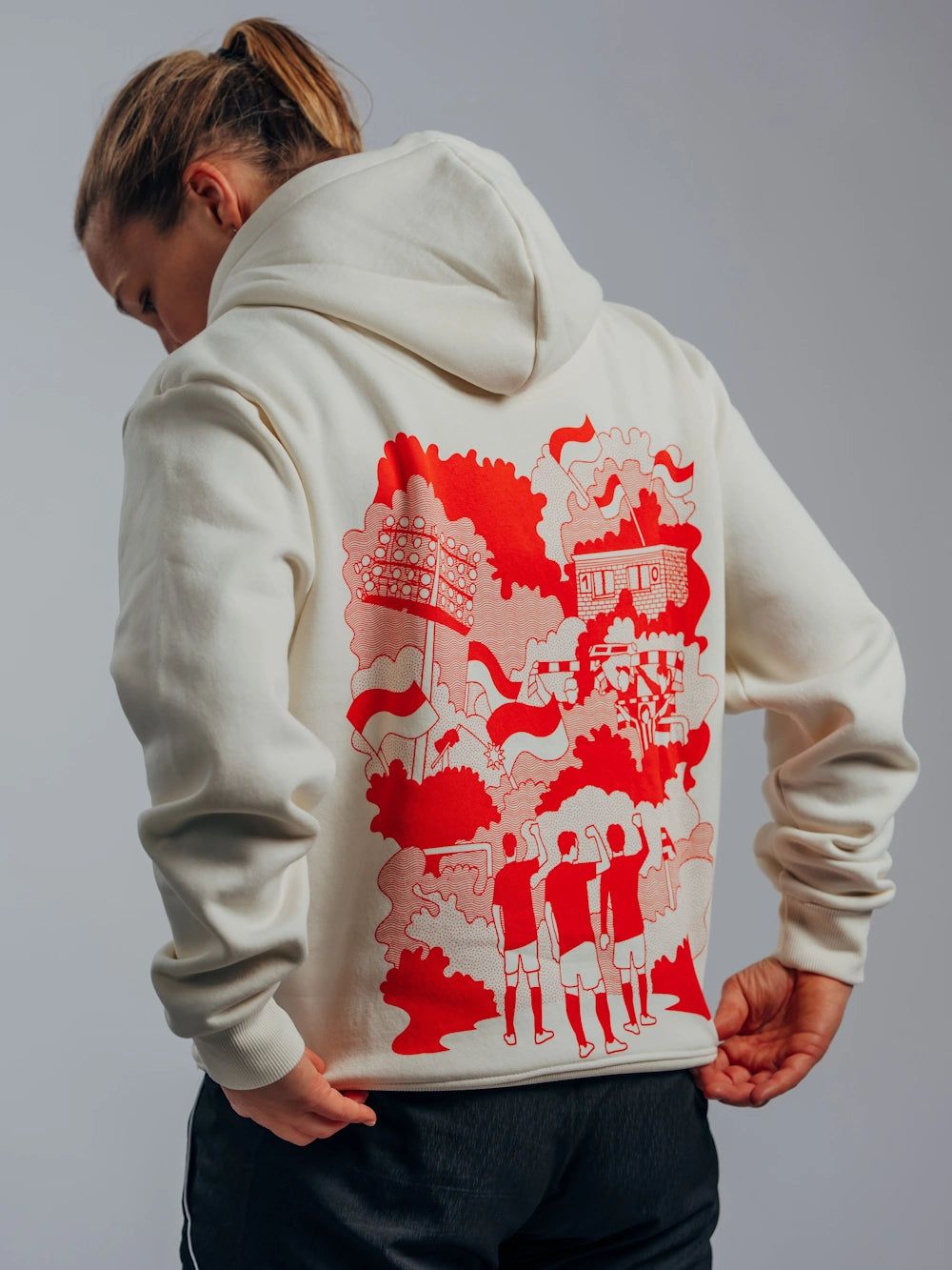 Eine Person trägt einen hellen Hoodie mit einem roten grafischen Druck auf dem Rücken, der drei Figuren und fantasievolle Landschaften zeigt.