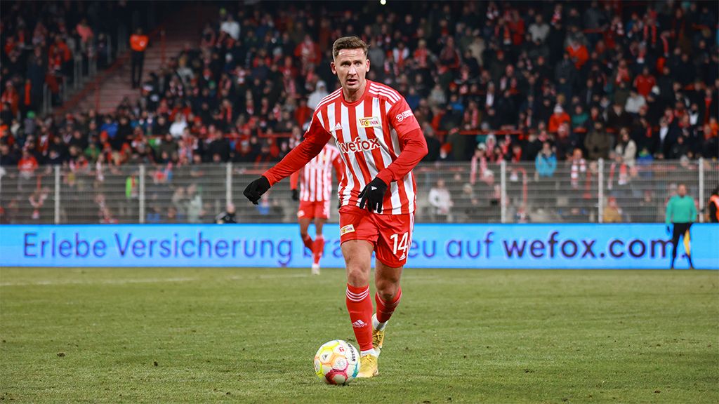 Spieler in rot-weißer Fußballausrüstung auf dem Platz, umgeben von jubelnden Fans im Stadion.