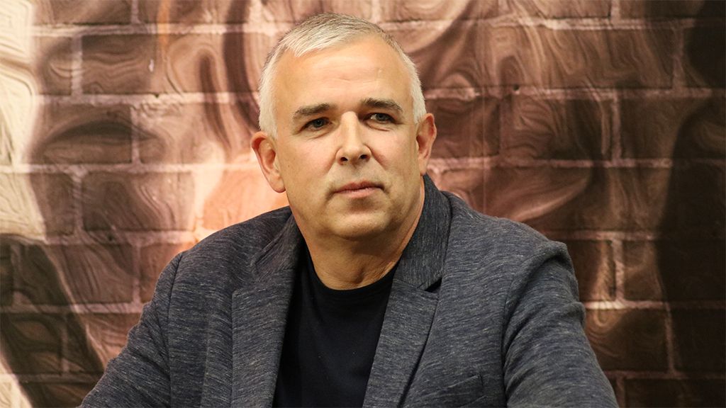 Ein Mann mit grauen Haaren sitzt vor einer gemusterten Wand und schaut nachdenklich in die Kamera.