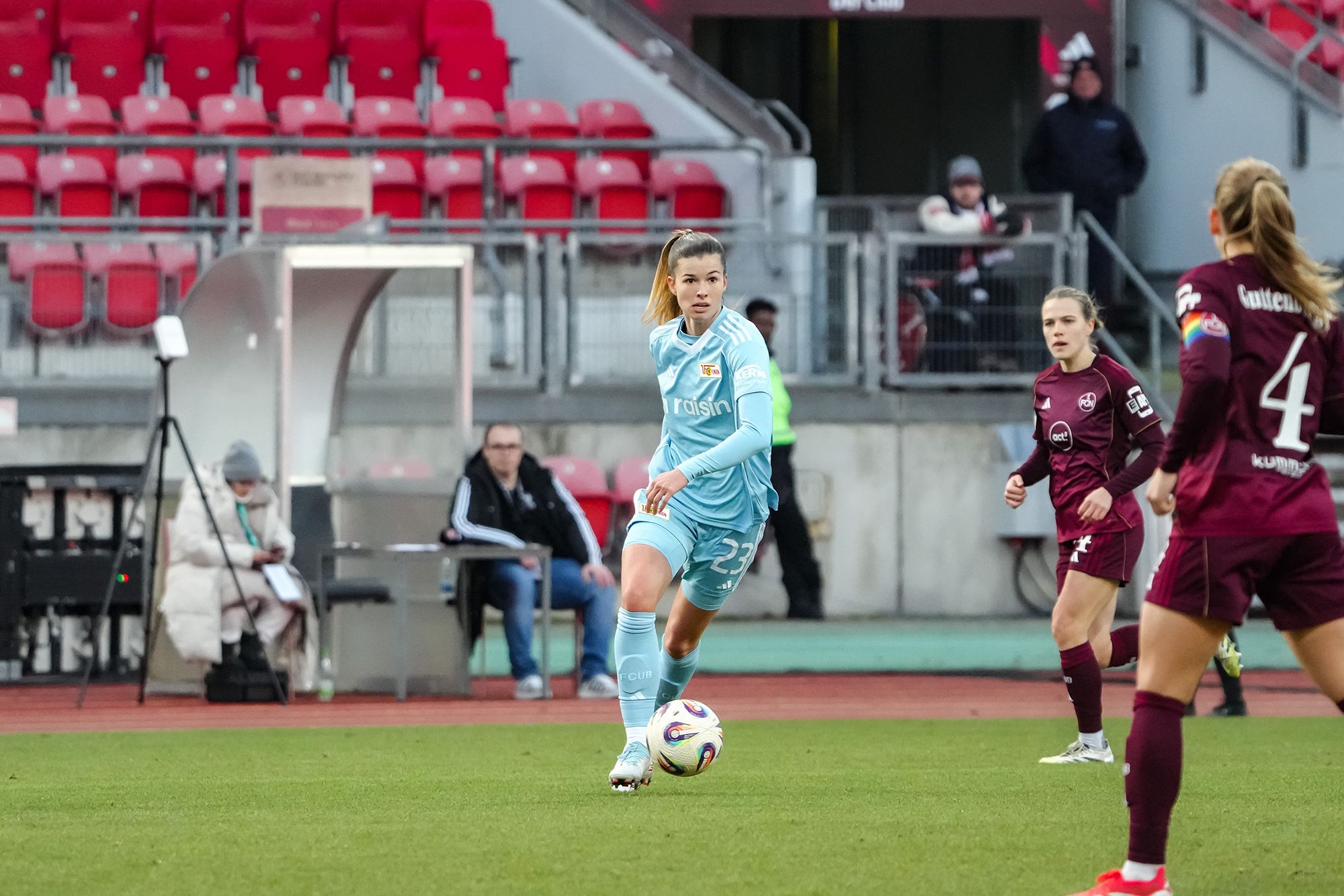 Eine Fußballspielerin in blauer Torwartkleidung steht auf dem Spielfeld und bereitet sich auf einen Pass vor. Zuschauer im Hintergrund.