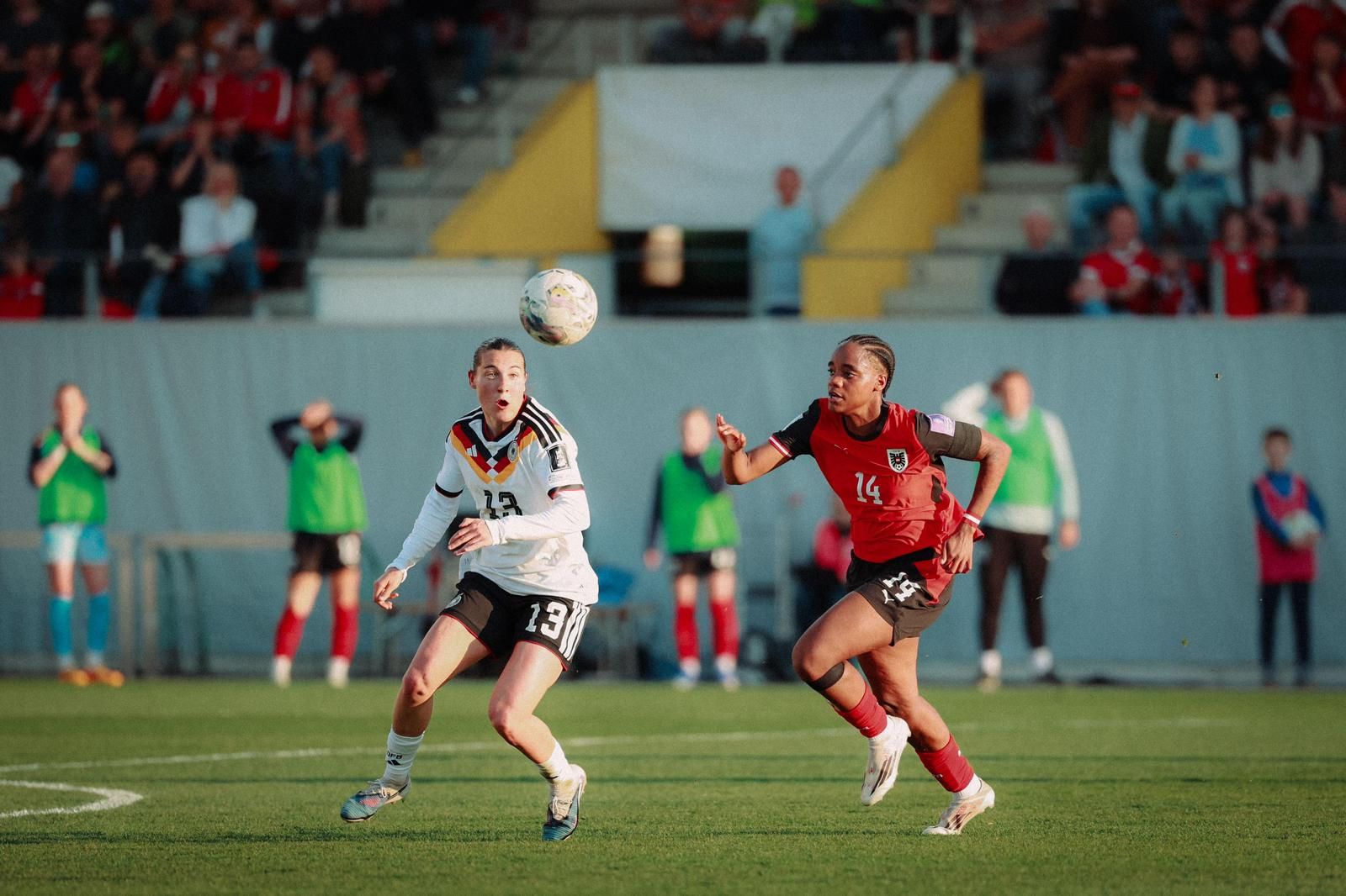 Zwei Fußballspielerinnen in action; eine spielt den Ball, während die andere sich verteidigt, im Hintergrund Zuschauer und Bank.